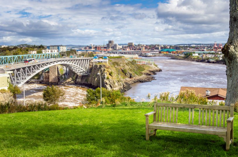 Photo of Saint John, New Brunswick, from a park near the Reversing Falls Bridge. An empty wooden bench is in Foreground.