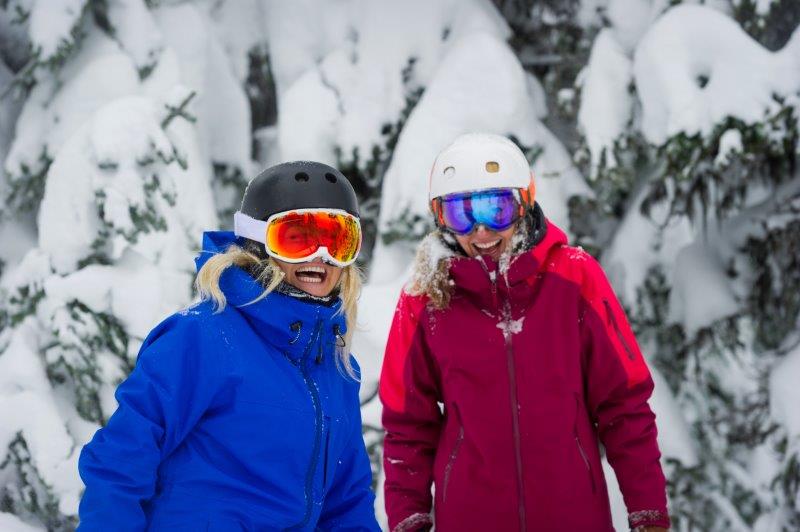 Twwo women in bightly colored ski jackets, goggles and ski helmets smiling.