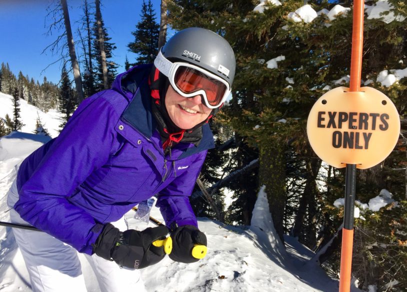 Woman in purple jacking smiling in front of Experts Only ski sign