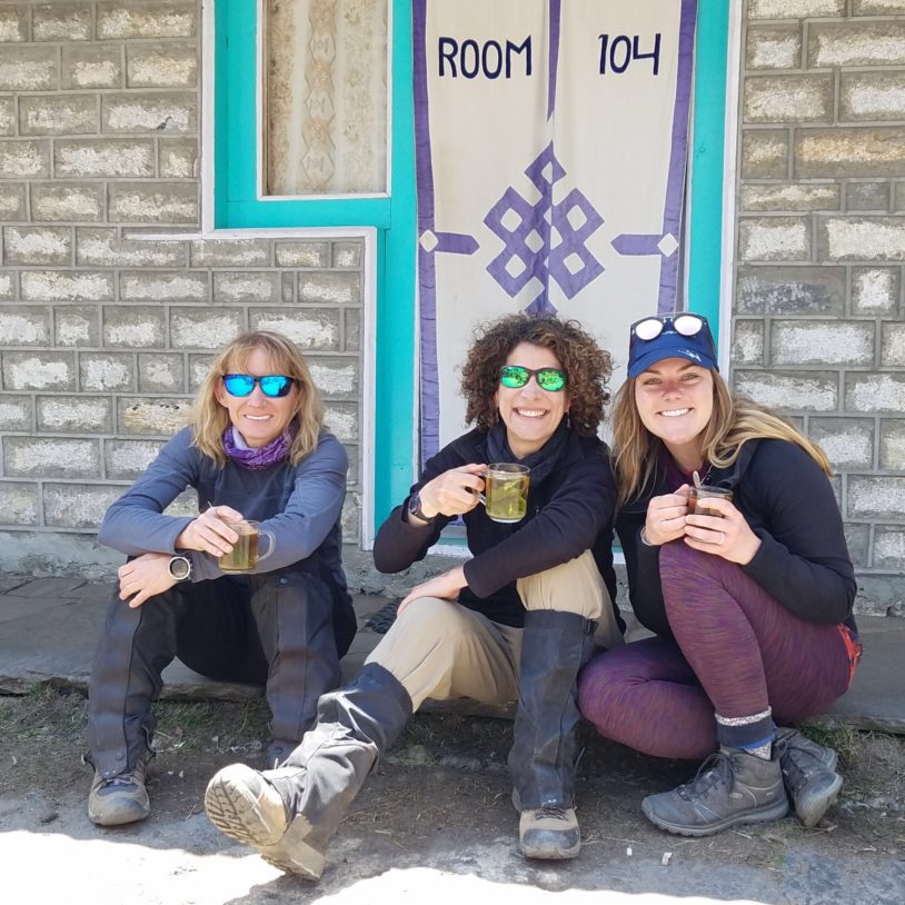 Three women laughing and smiling sitting against a building before hiking.